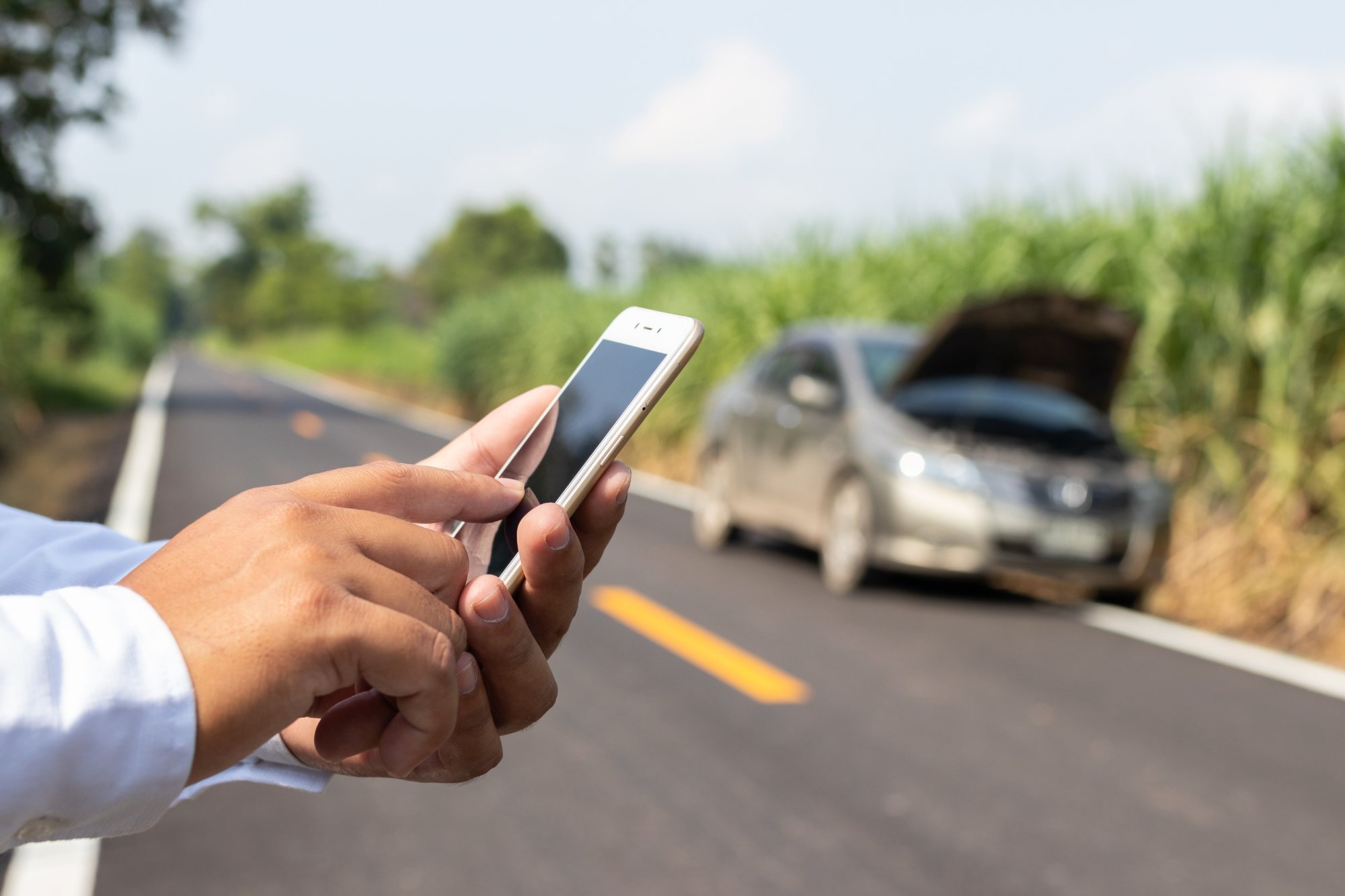 Person holding smartphone near damaged car on roadside with rural landscape