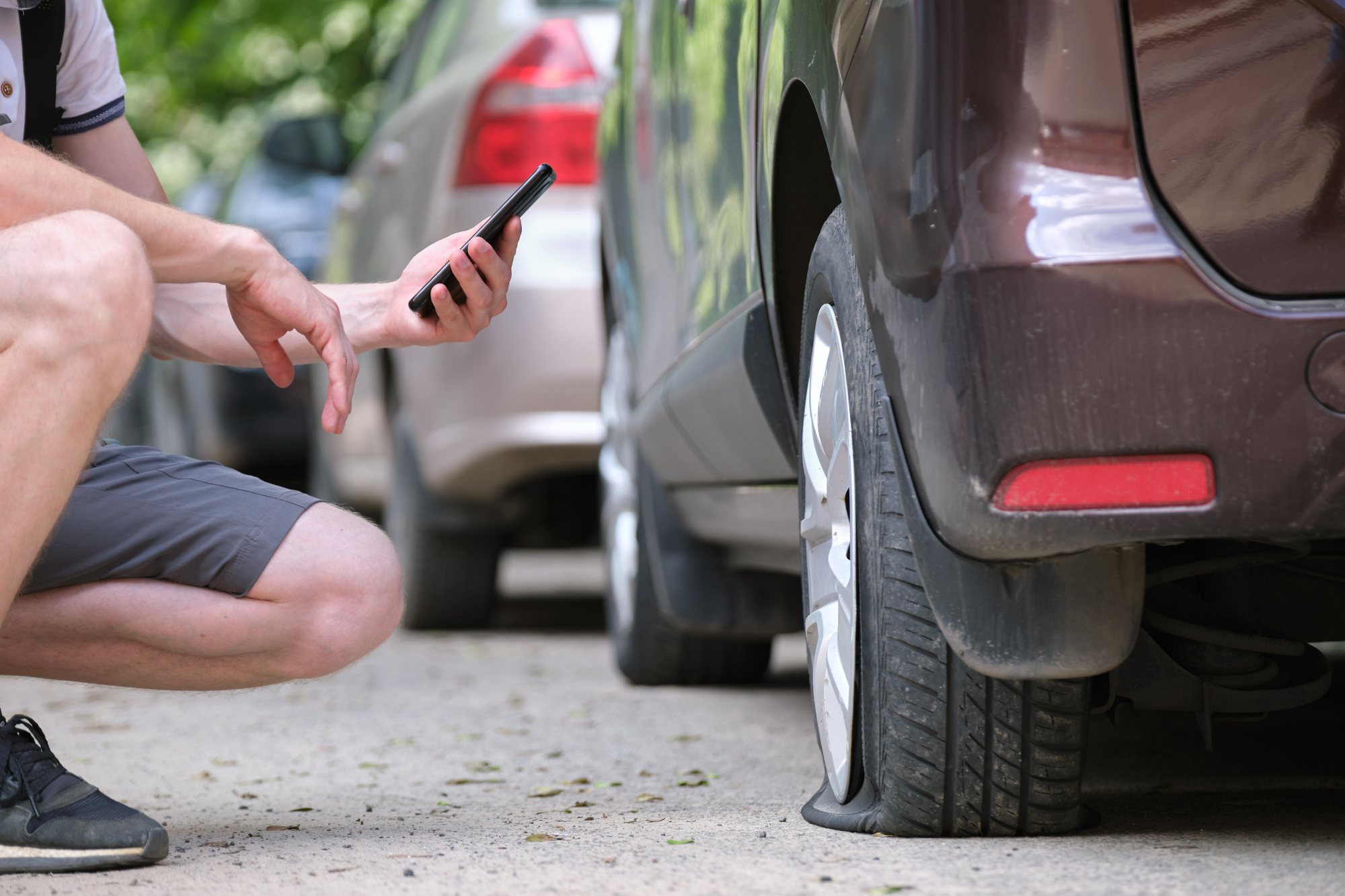 Man holding smartphone while inspecting parked car tire and wheel