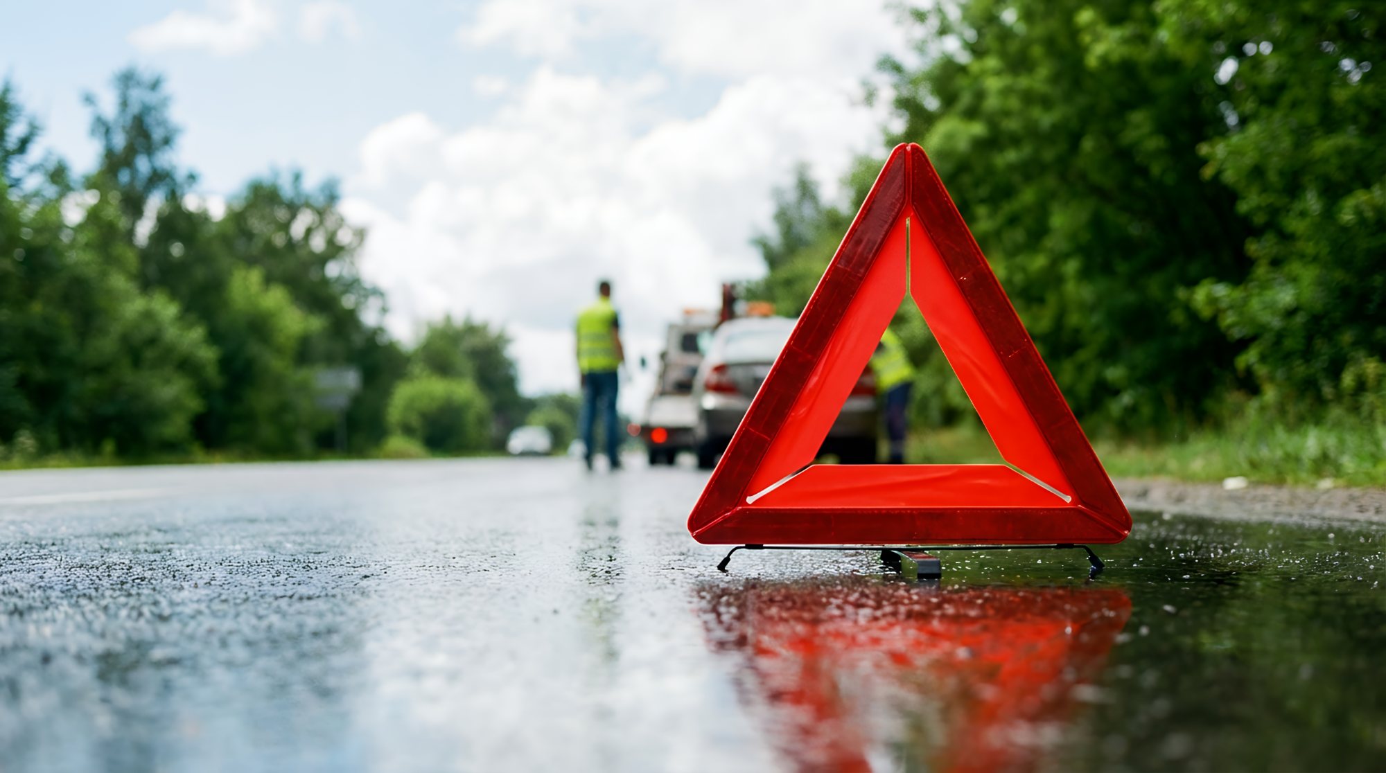 Red warning triangle on wet road with emergency vehicles and personnel in background