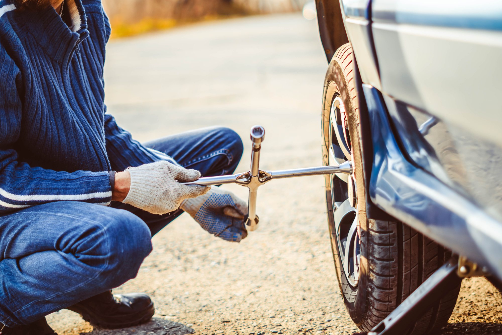Person in blue sweater using lug wrench to change car tire on road